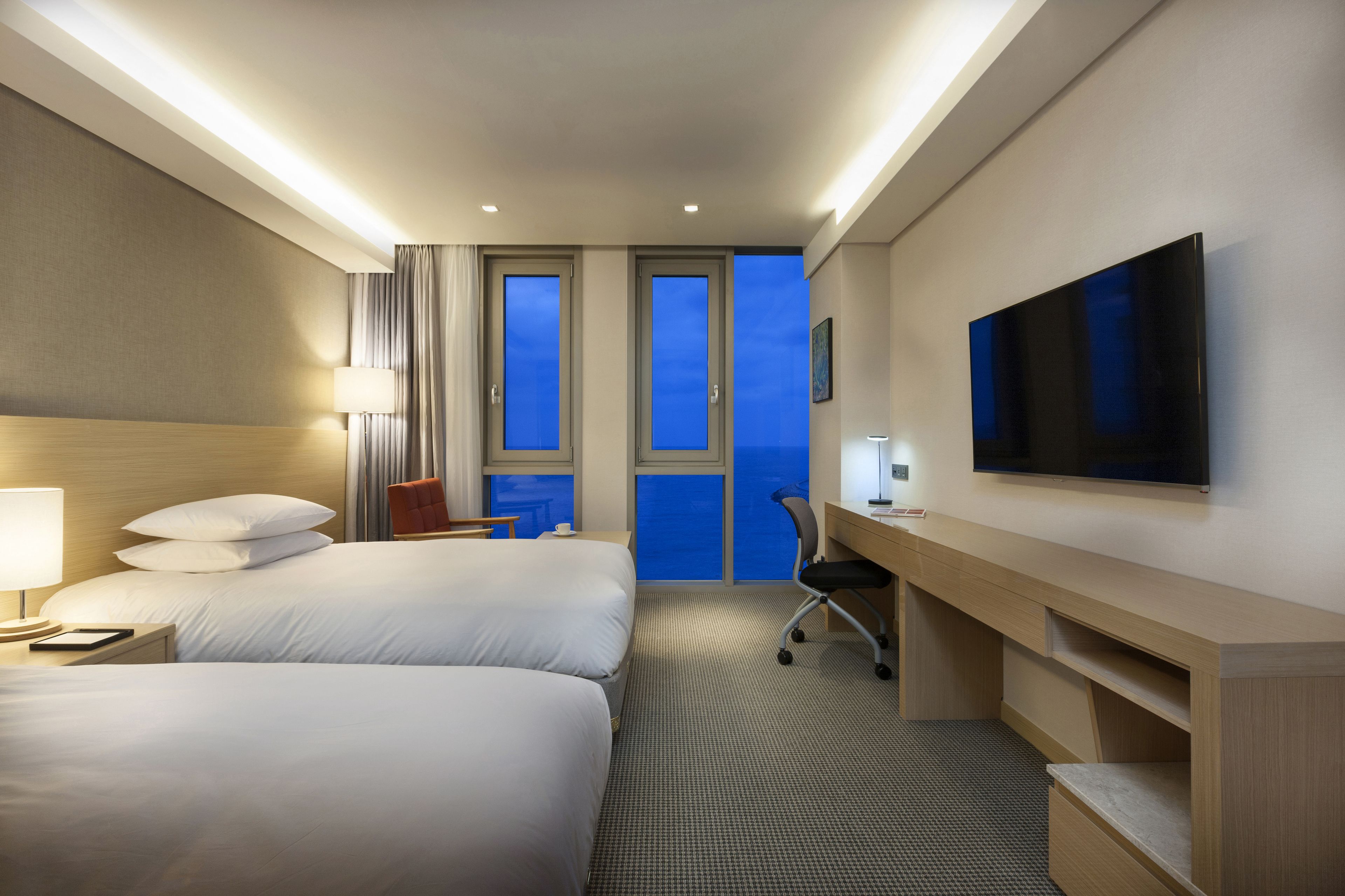 A hotel room with two beds, a desk, a chair, a TV, and a view of the sky through the windows.