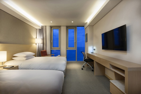 Habitación de hotel con dos camas, un escritorio, una silla, un televisor y vistas al cielo a través de las ventanas.
