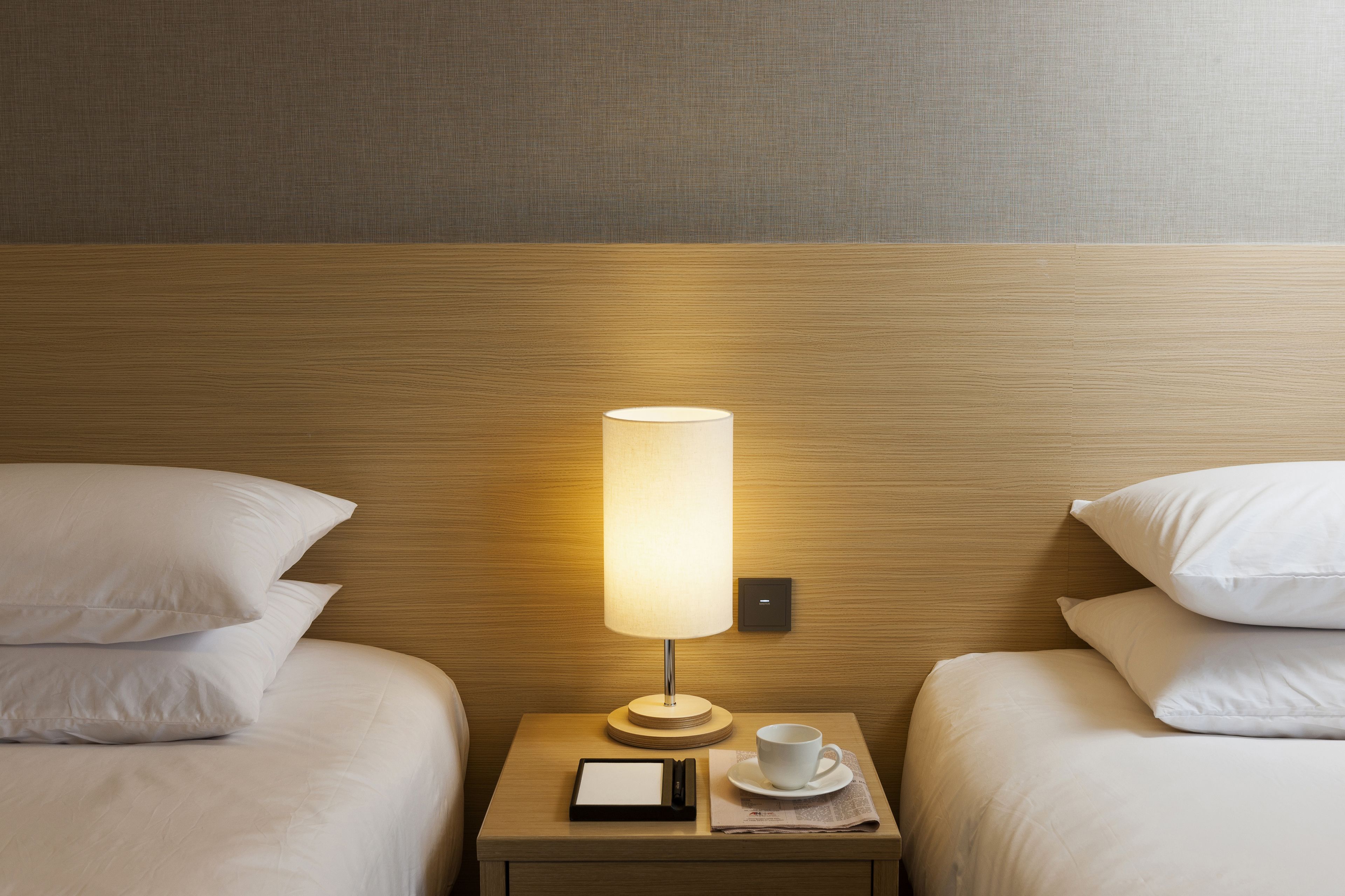 A neatly made bed with white linens, a bedside table with a lamp, a cup, and a menu.