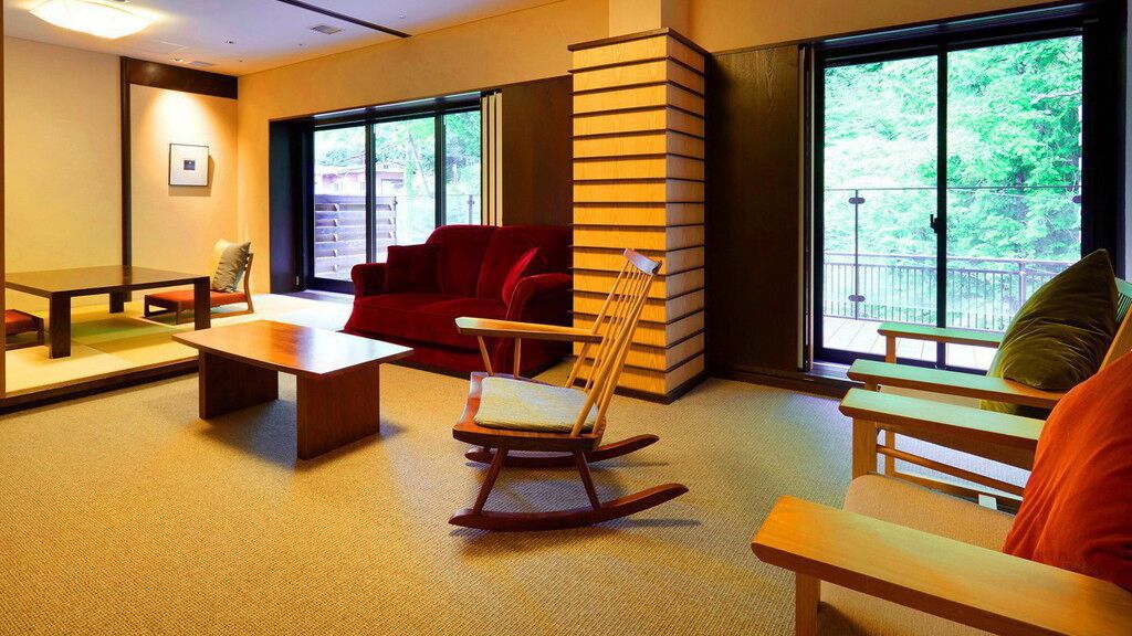 Ein Zimmer mit einem roten Sofa, einem Schaukelstuhl, einem Holztisch und großen Fenstern mit Blick ins Grüne.