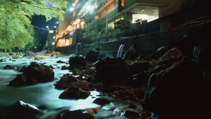 Un edificio de varios pisos con ventanas y balcones iluminados, ubicado junto a un río con rocas y árboles.