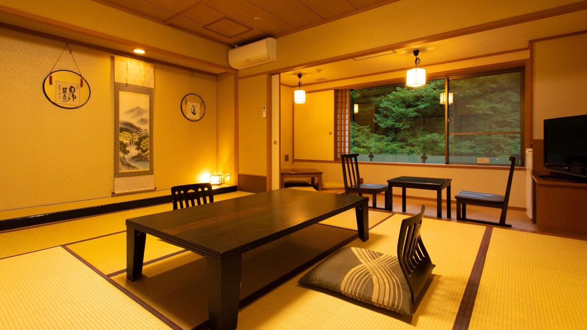 A traditional Japanese room with tatami flooring, a low table, chairs, a television, and a view of greenery outside.