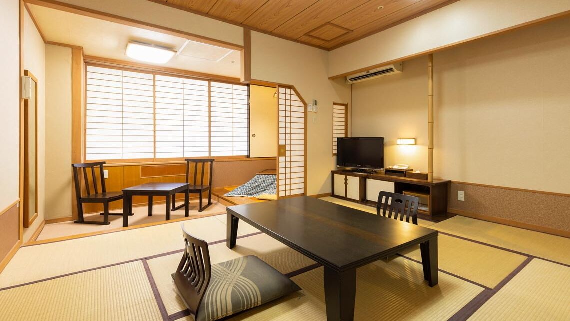 A traditional Japanese room with tatami flooring, a low table, chairs, a television, and a sliding door.