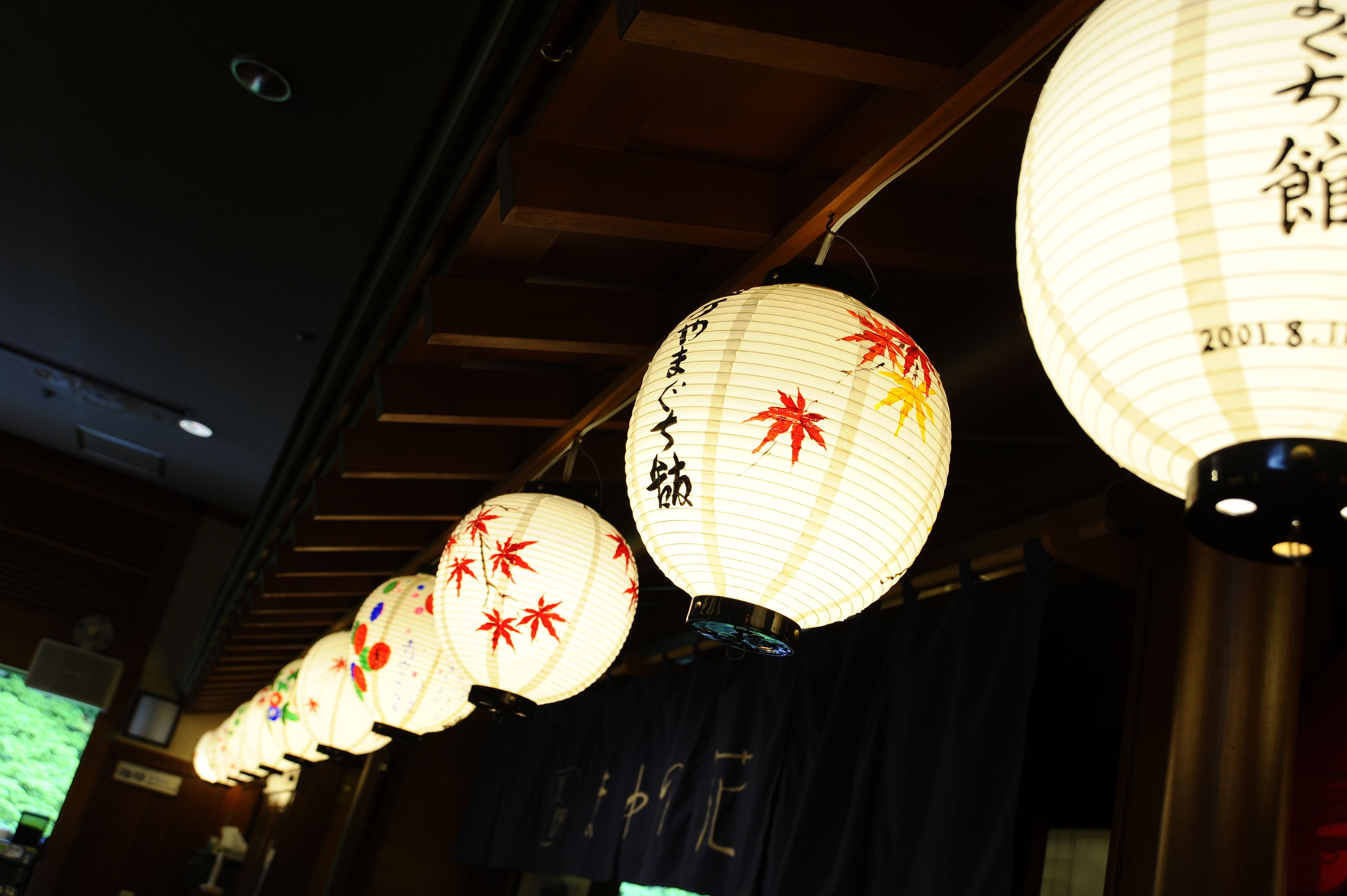 Une rangée de lanternes japonaises traditionnelles en papier, ornées de feuilles d’érable rouges et de calligraphies, est suspendue au plafond.