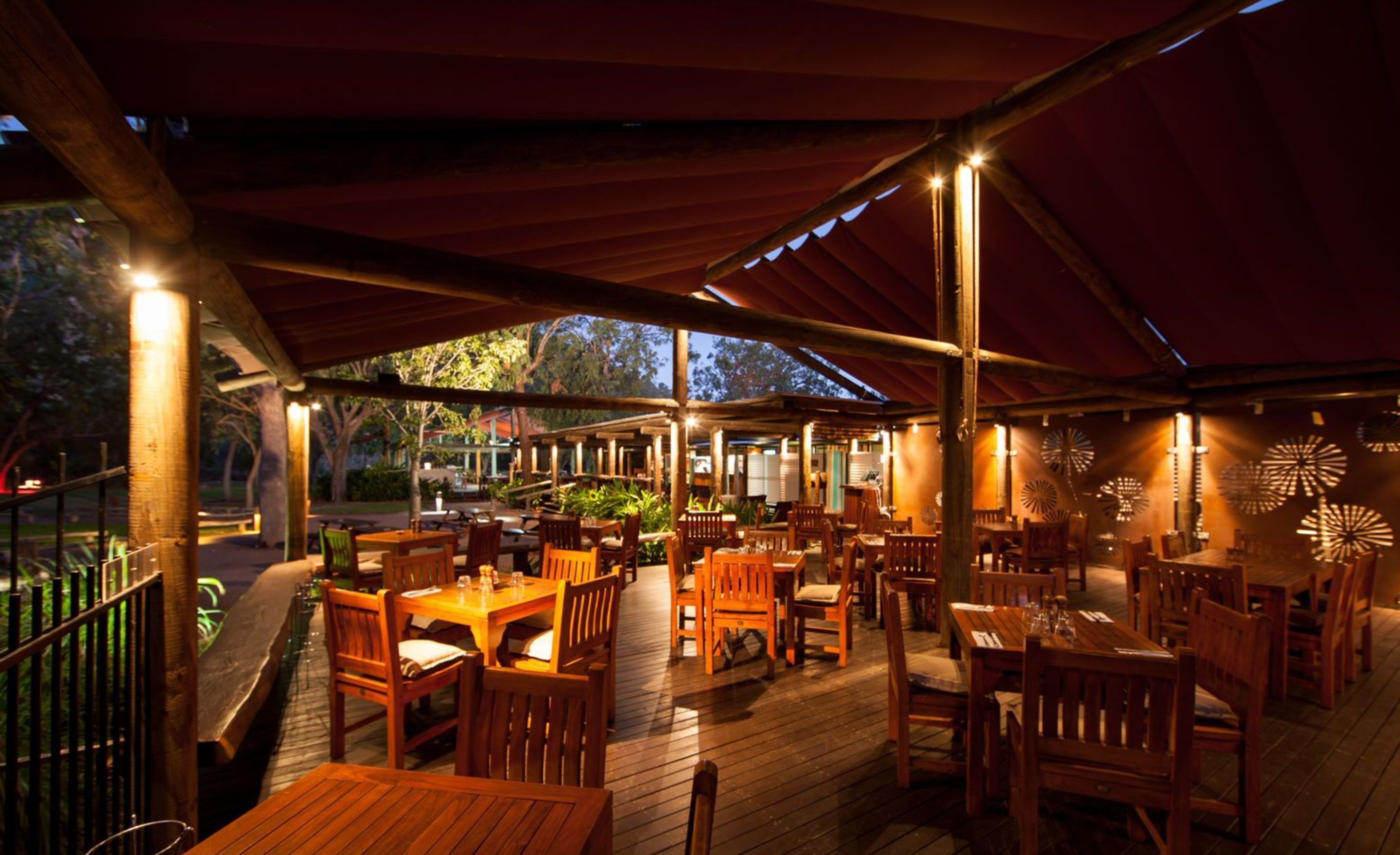 An outdoor dining area with wooden tables and chairs, covered by a red canopy, and surrounded by trees.