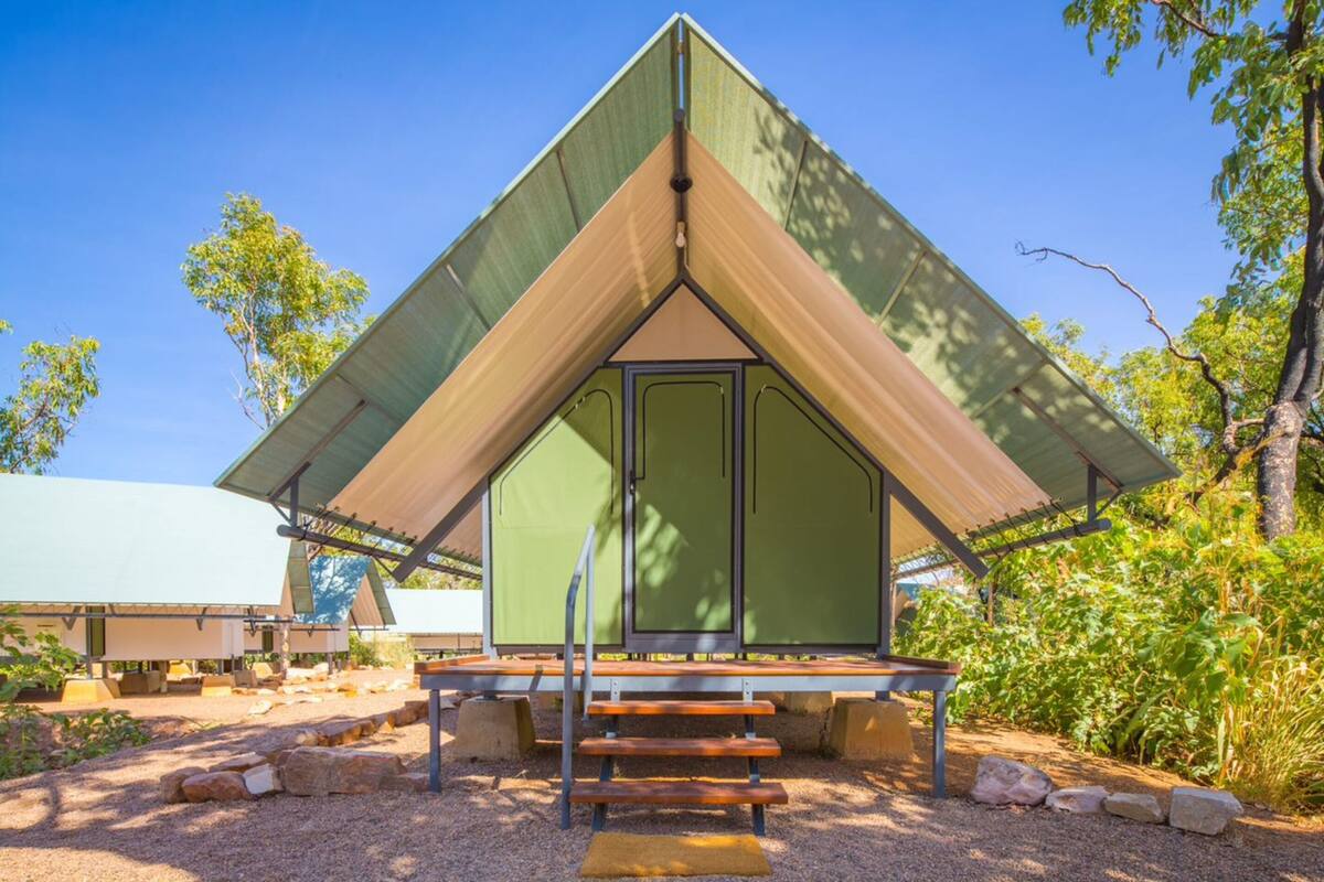 A triangular tent-like structure with a bench in front, surrounded by gravel and rocks, with trees in the background.