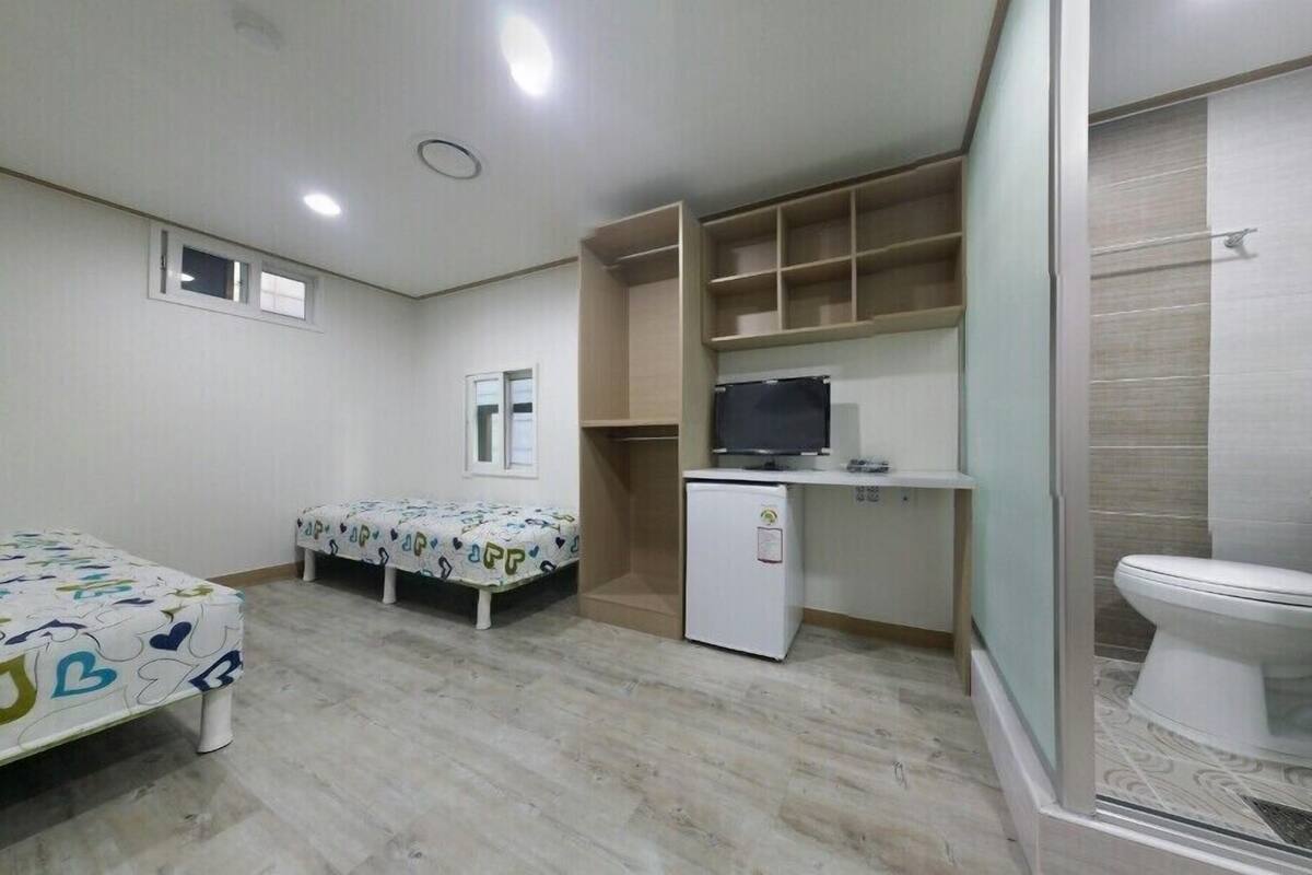 A small, clean room with a bed, a desk with a computer, a toilet, and a shower area.