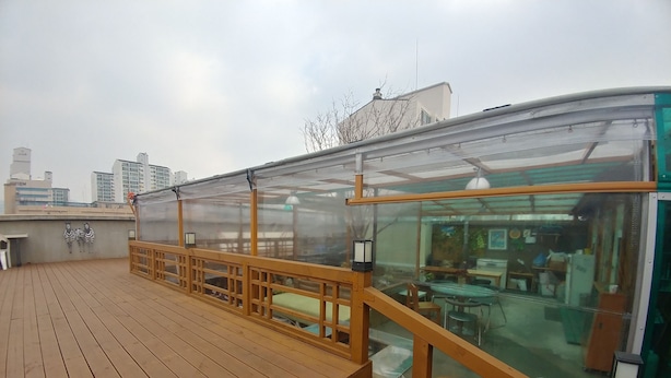 A rooftop terrace with wooden flooring, a glass enclosure, and a seating area with stools. - Don Quixote Stay (Seoul)