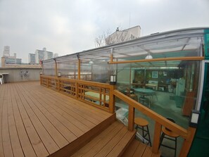 Een terras op het dak met houten vloer, een glazen omheining en een zithoek met krukjes.