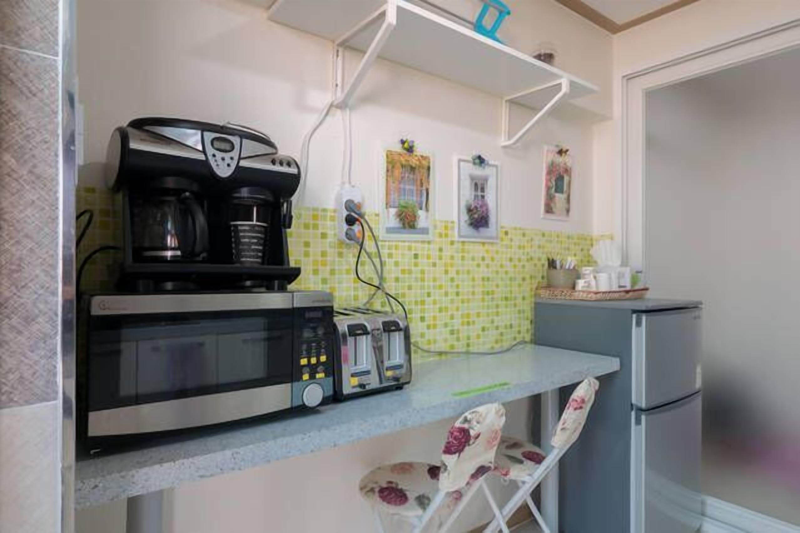 A compact kitchen with a coffee maker, toaster, and microwave. There is a small refrigerator and a chair with a floral pattern.