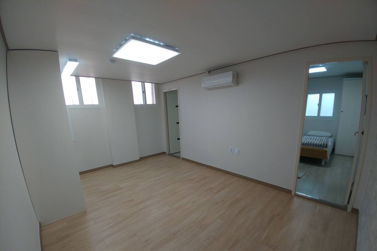 A spacious room with a wooden floor, a ceiling light, an air conditioning unit, and a door leading to another room with a bed.