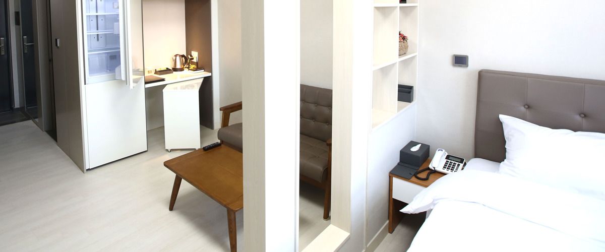 A hotel room with a bed, bedside table with a phone and a lamp, a desk with a chair, and a wardrobe.
