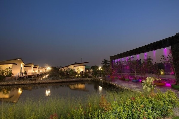 A nighttime view of a resort with a pool, illuminated buildings, and a landscaped area with plants.
