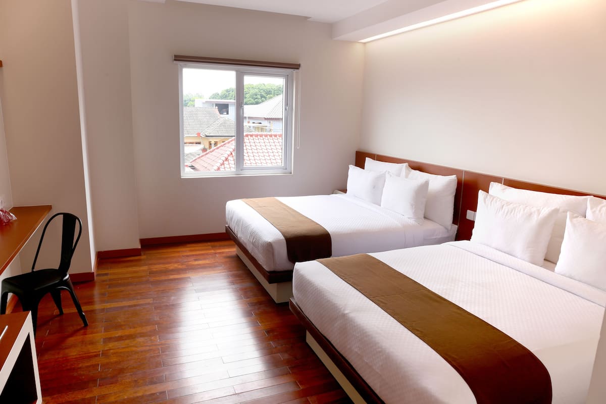 Een hotelkamer met twee bedden, een houten vloer, een raam met uitzicht op gebouwen, en een bureau met een stoel.