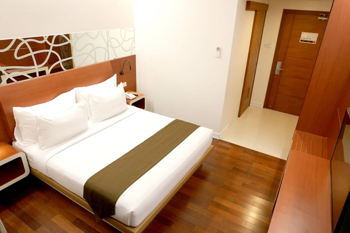 Een net opgemaakt bed met wit beddengoed en een bruine accentkleur, een houten nachtkastje met een telefoon en oplader, en een houten vloer.