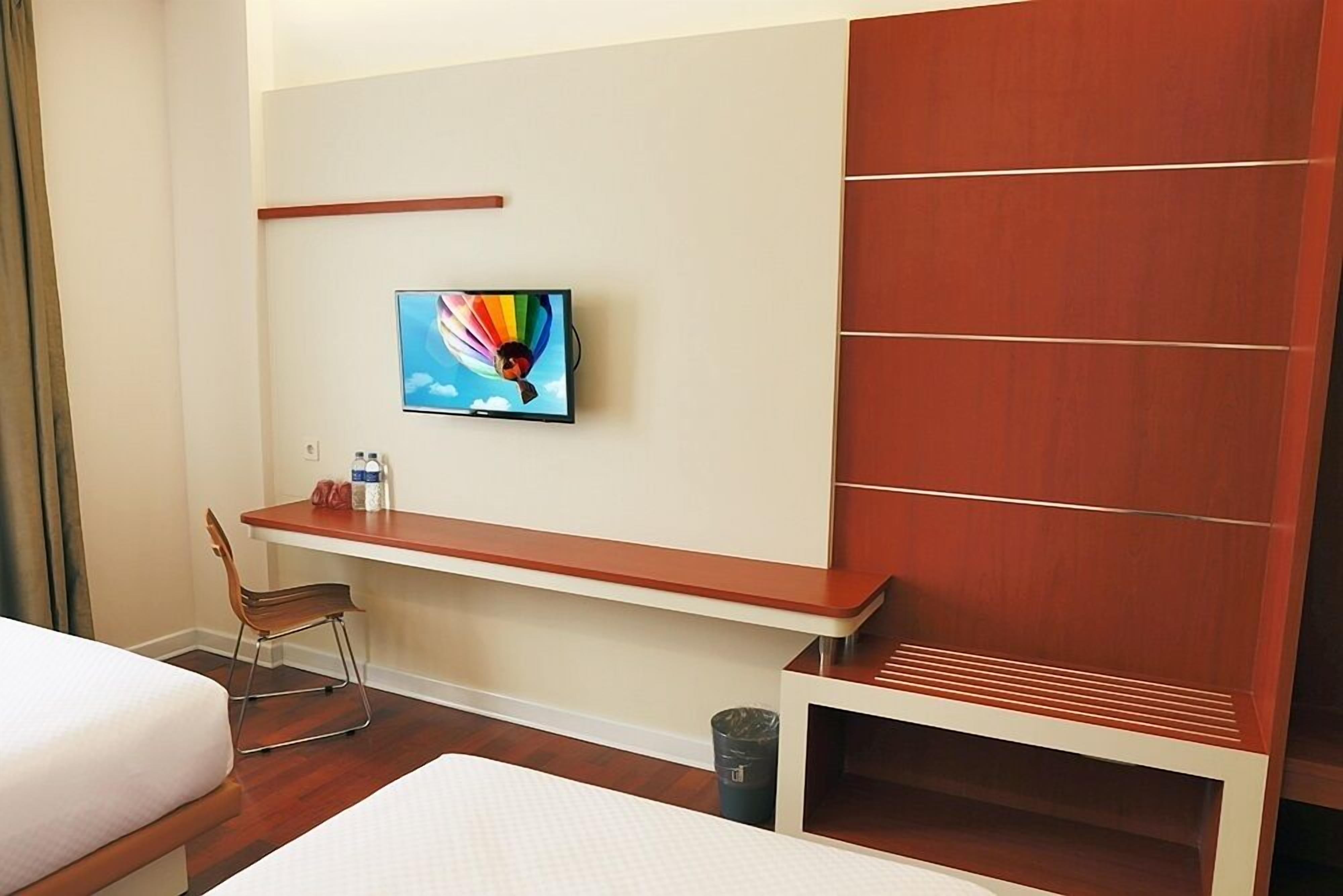 Habitación de hotel con cama, escritorio, silla, televisor montado en la pared y un cubo de basura.