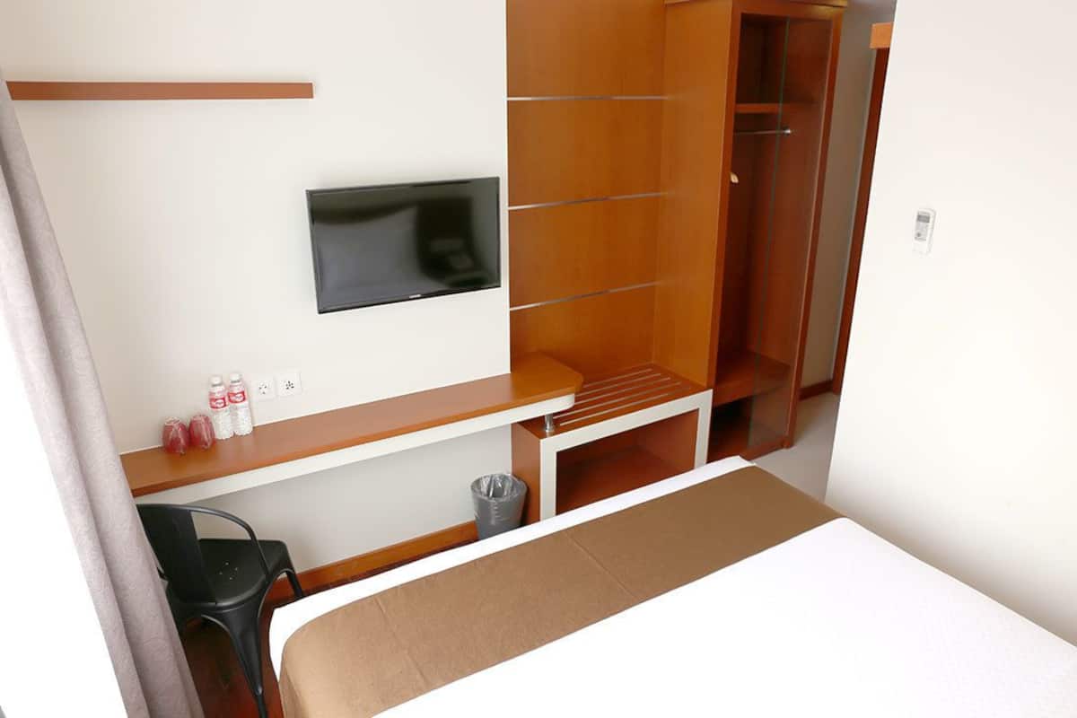Een hotelkamer met een bed, een aan de muur bevestigde televisie, een houten garderobe, een klein tafeltje met flessen en een prullenbak.