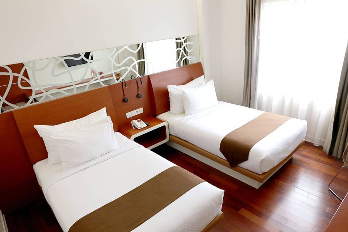 Een hotelkamer met twee bedden, een houten hoofdbord, een nachtkastje met een telefoon en een raam met gordijnen.