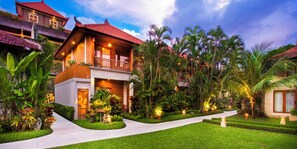Una casa de dos pisos con un balcón, rodeada de abundante follaje y palmeras.