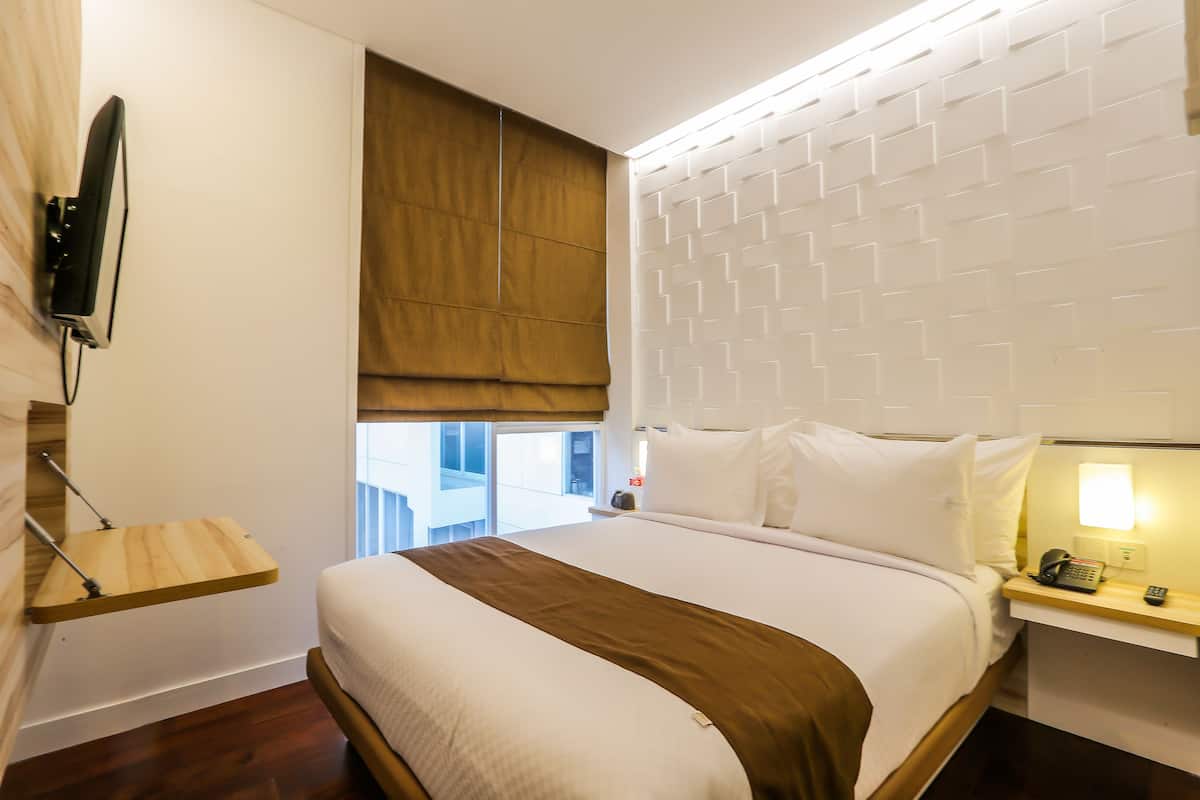 A hotel room with a bed, a bedside table with a lamp, a mounted TV, and a window with a view of the outside.