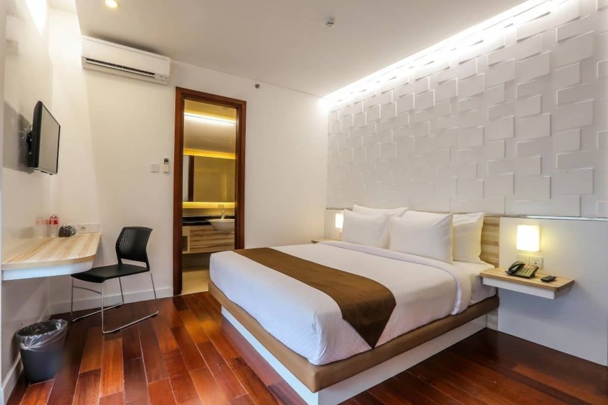 A modern hotel room with a large bed, a bedside table with a lamp, a chair, and a TV.