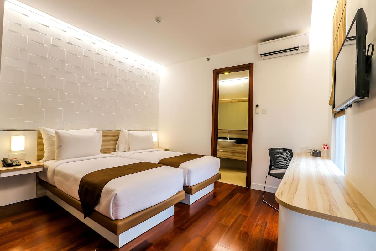 A hotel room with a double bed, bedside tables, a small desk, a TV mounted on the wall, and an air conditioning unit.