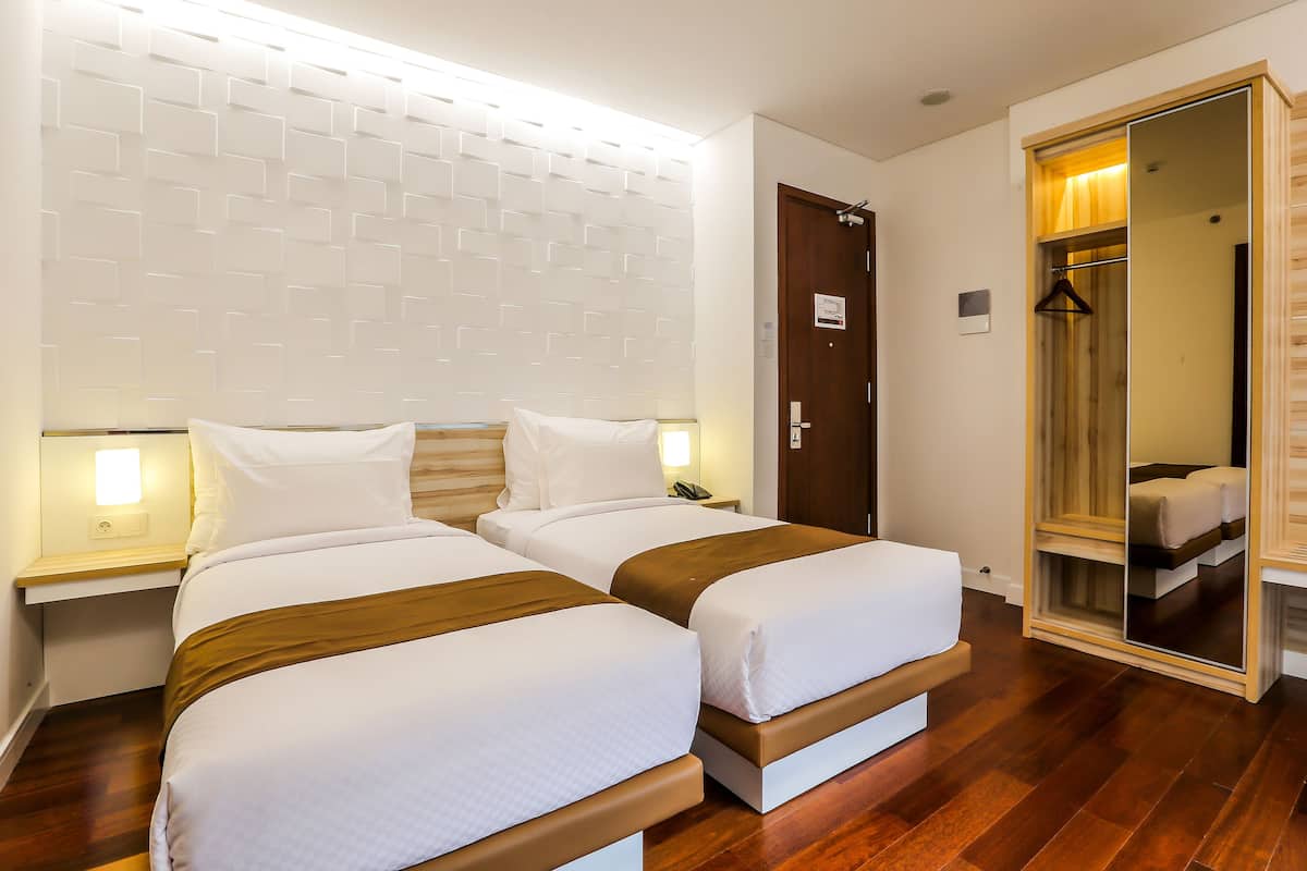 A hotel room with two beds, wooden headboard, bedside tables with lamps, a wooden floor, and a large mirror.