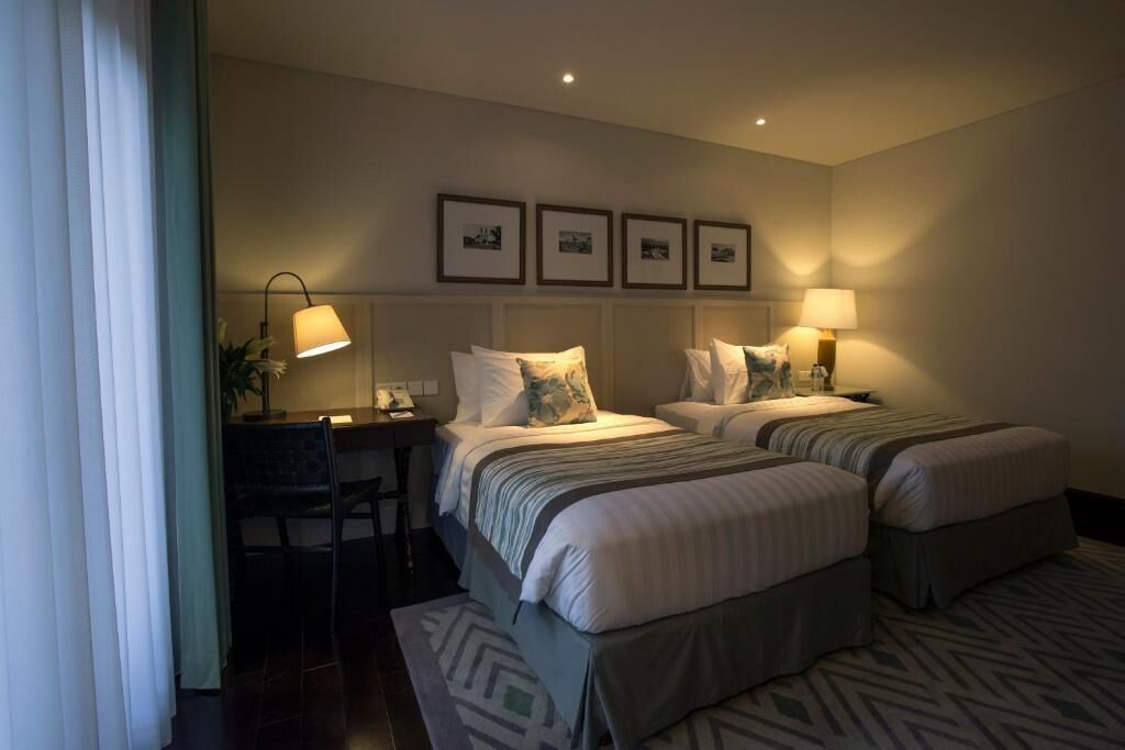 Habitación de hotel con dos camas, un escritorio y una silla. Hay cuadros en la pared y lámparas en las mesitas de noche.