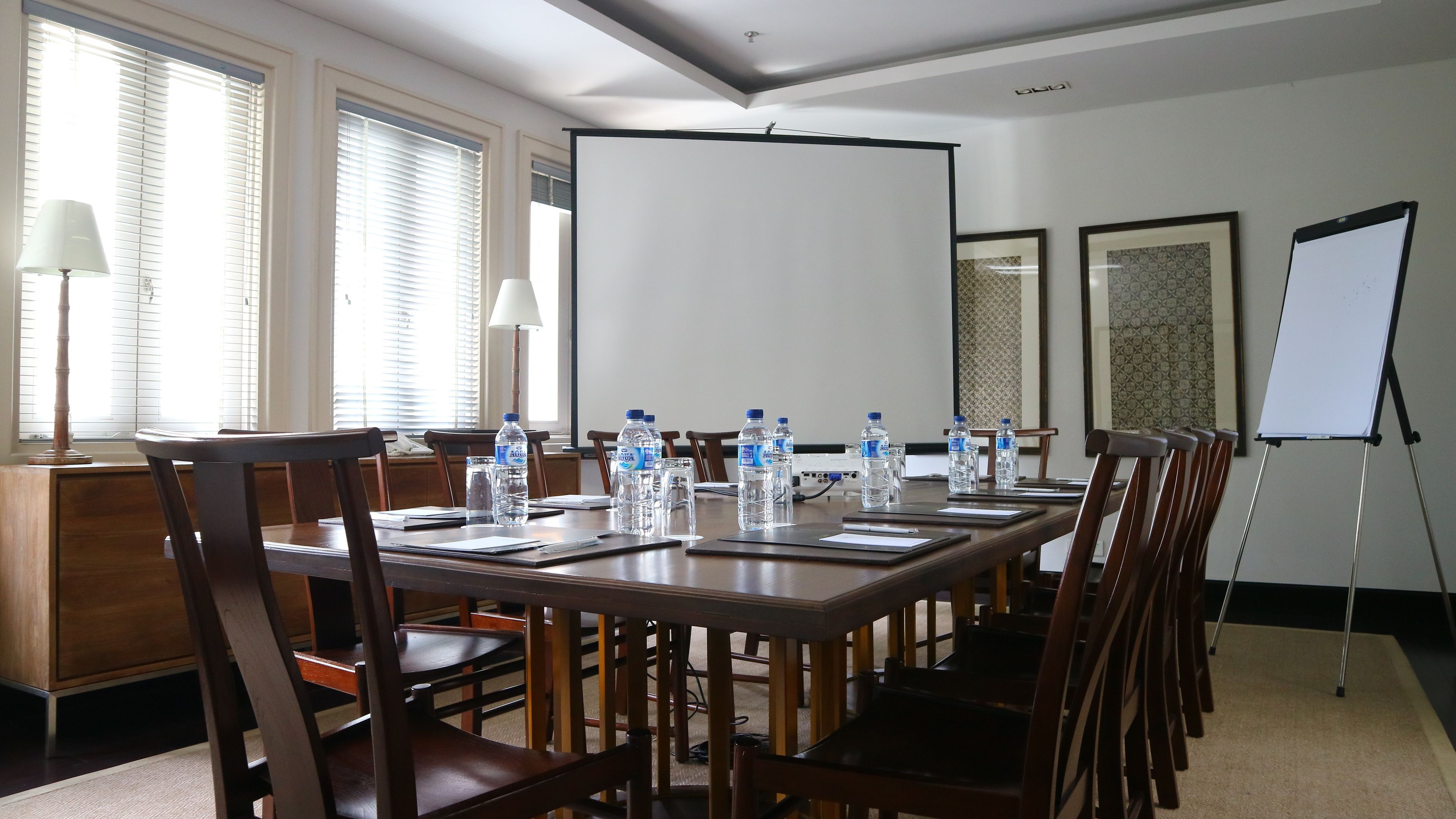 Un salón de conferencias con una larga mesa, sillas, botellas de agua, una pantalla de proyector y una pizarra.