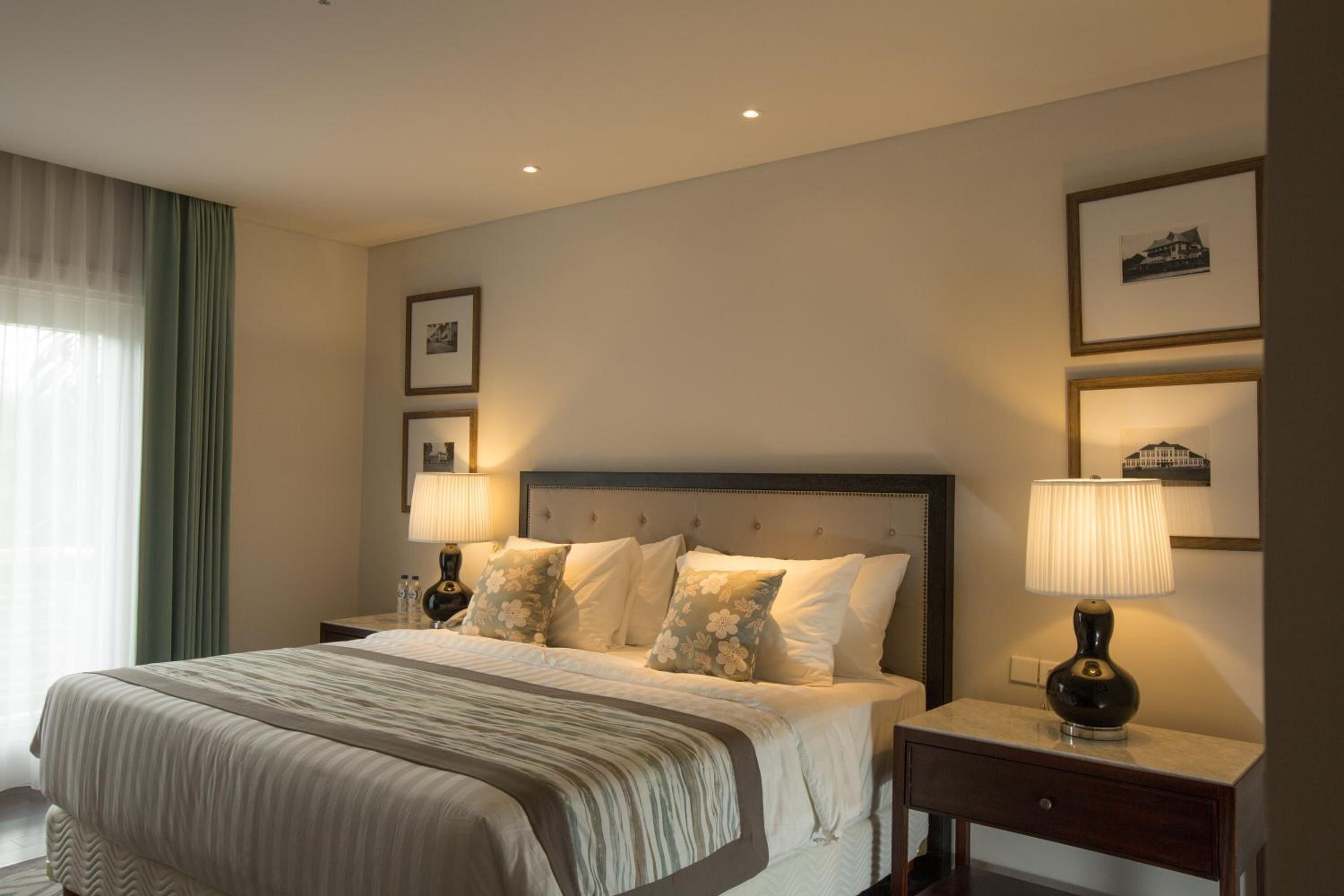 A hotel room with a large bed, two bedside tables with lamps, a nightstand, and a wall-mounted shelf with framed pictures.