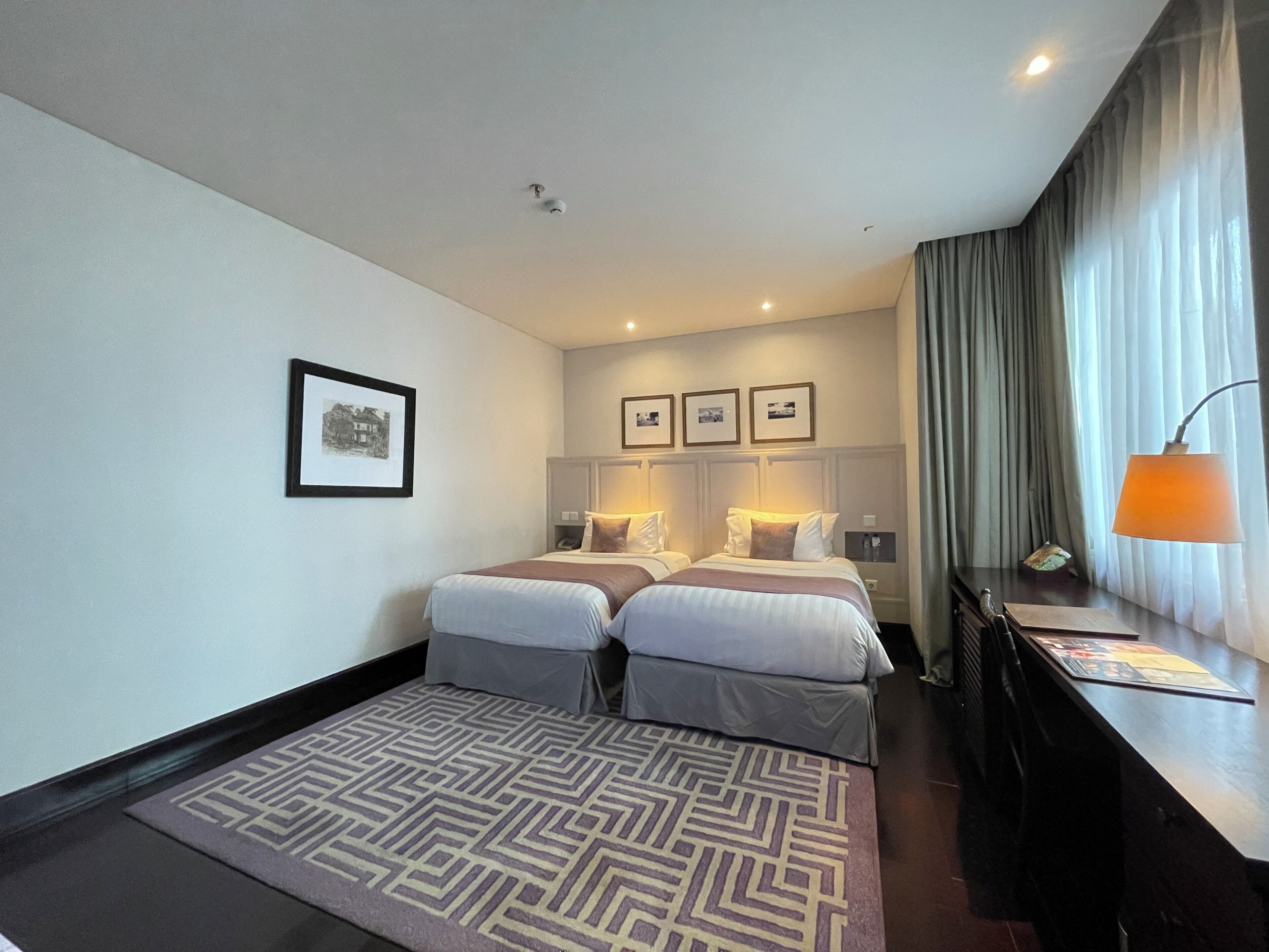 A hotel room with two beds, a desk, a lamp, and framed pictures on the wall.