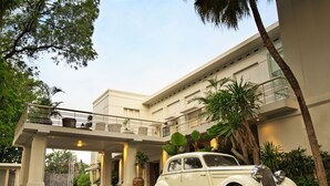 Une voiture blanche, modèle classique, est garée devant un bâtiment moderne doté d’un balcon et entouré d’une végétation luxuriante.