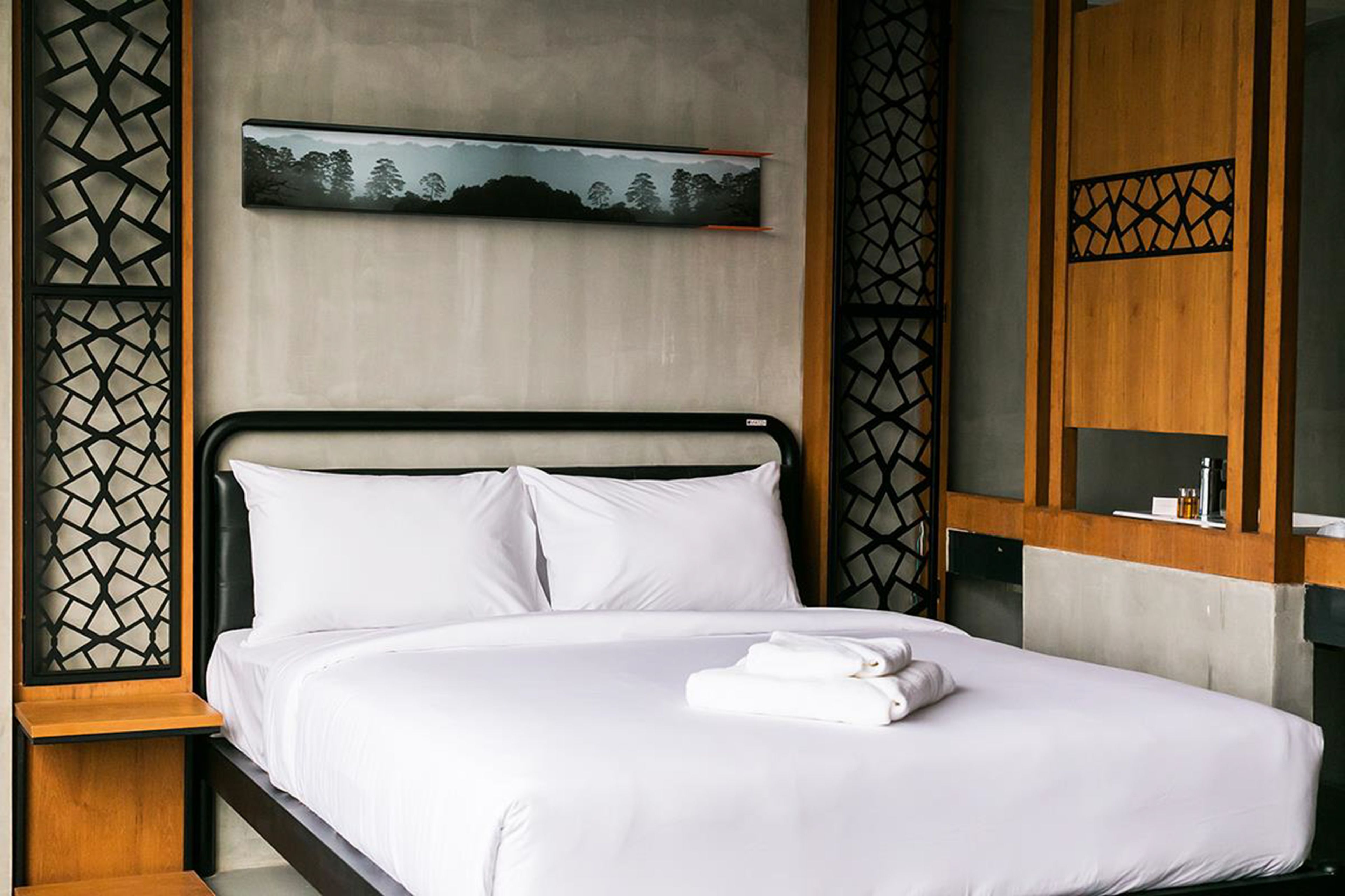 A neatly made bed with white linens and a folded towel, a wall-mounted artwork depicting a landscape, and a wooden nightstand with a small table.
