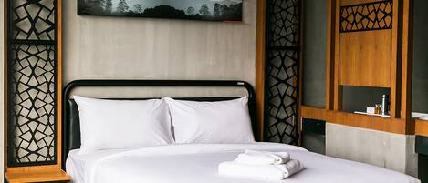 A neatly made bed with white linens and a folded towel, a wall-mounted artwork depicting a landscape, and a wooden nightstand with a small table.
