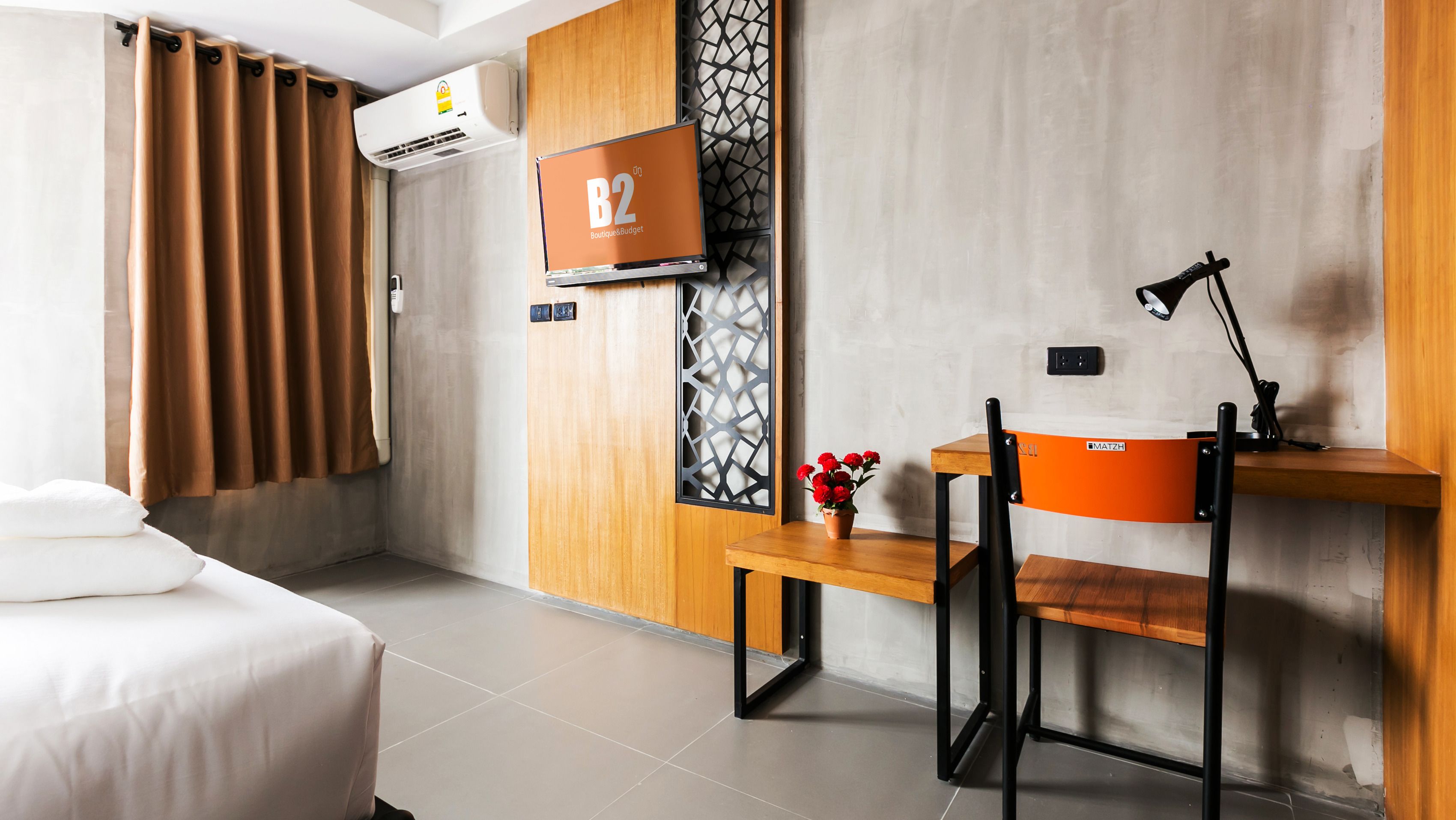 A modern hotel room with a bed, a desk with a chair, a TV, and an air conditioning unit.