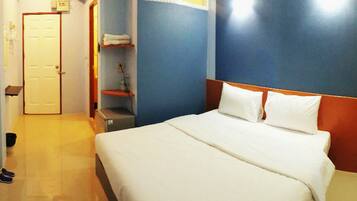 A hotel room with a bed, a door, a shelf with folded towels, and a small desk with a computer.