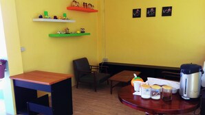 A room with a wooden table, a chair, a bench, and a coffee machine. There are shelves with figurines and a wall with three pictures.