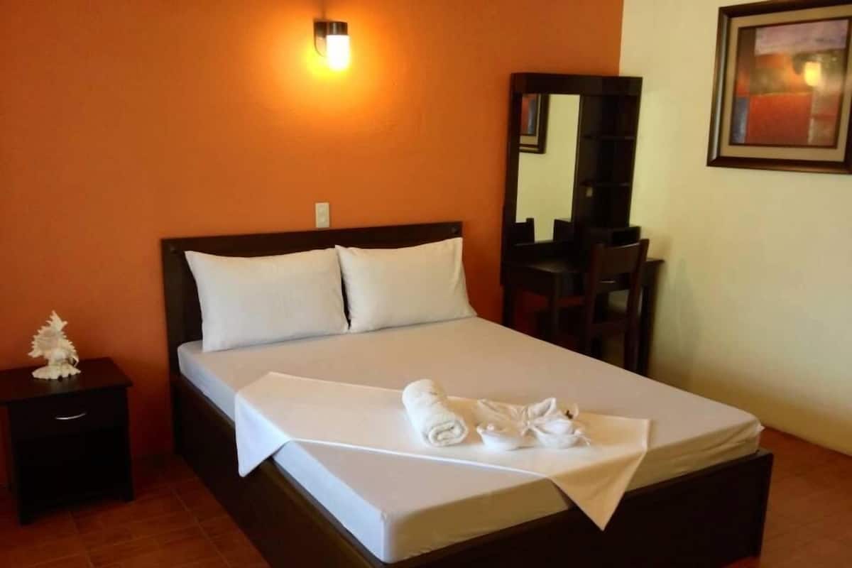 A neatly made bed with white linens and towels, a nightstand with a decorative item, a desk with a chair, and a wall-mounted mirror.
