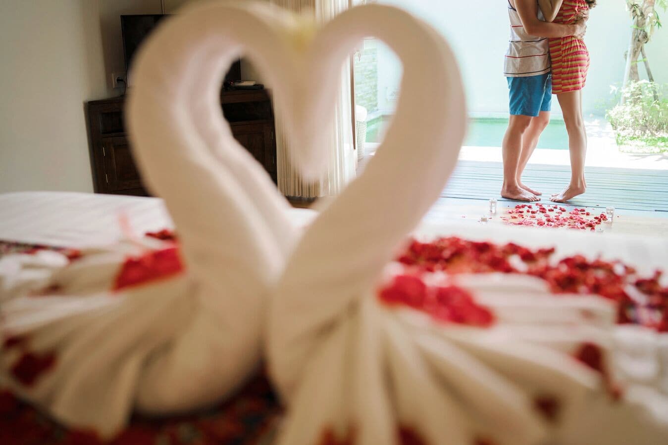 Una decoración en forma de corazón hecha de material blanco sobre una cama con pétalos rojos, un armario de madera y una persona al fondo.