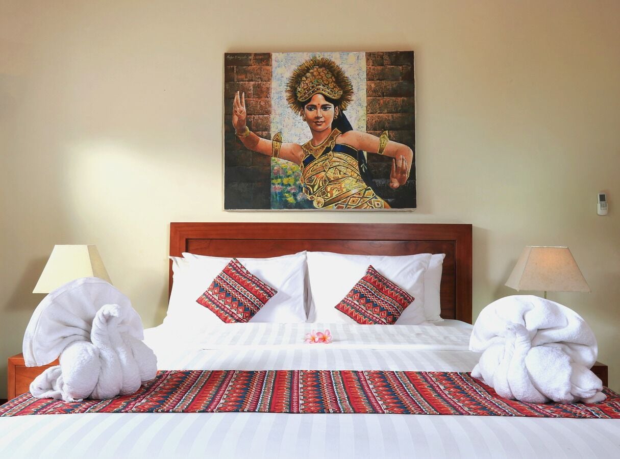 A hotel room with a bed, pillows, a painting on the wall, and a lamp on the nightstand.