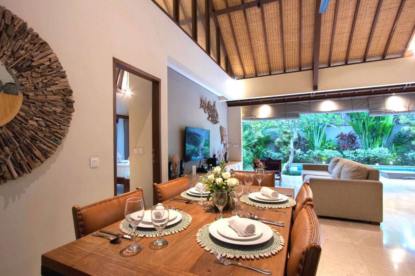 A dining area with a wooden table set for four, a television in the background, and a view of a lush garden through large windows.