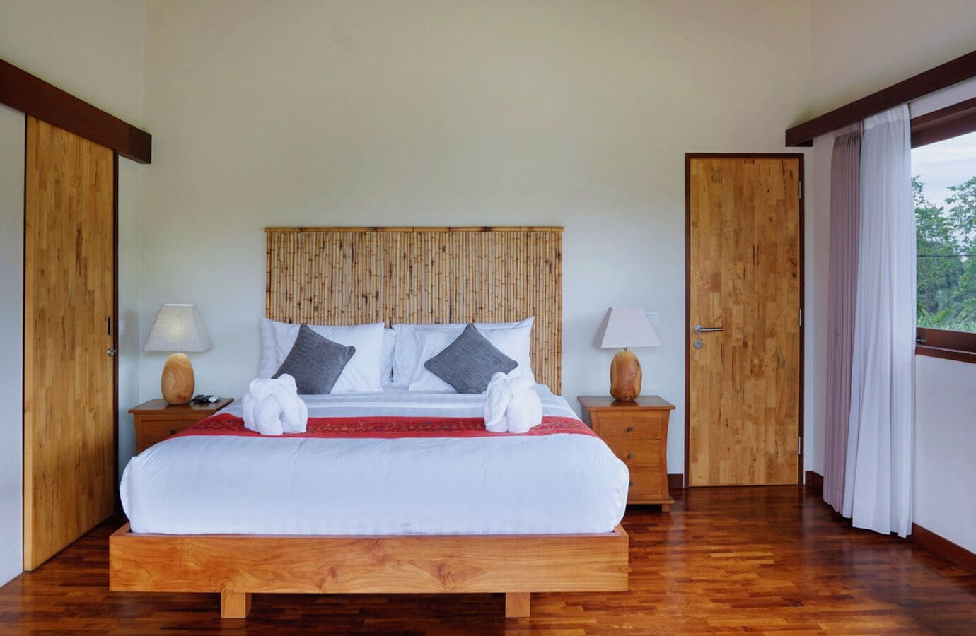 A large bed with a wooden headboard, two bedside tables with lamps, and a view of the outdoors through a large window.