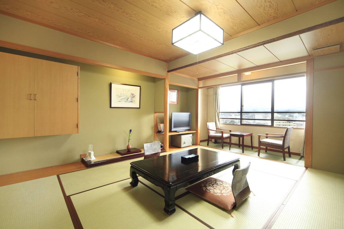 A traditional Japanese room with tatami flooring, a low table, a television, and a view of the city through large windows.