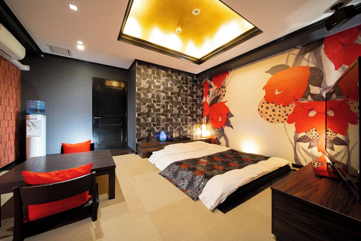 A modern hotel room with a bed, a desk, and a chair. There is a water dispenser and a wall-mounted heater.