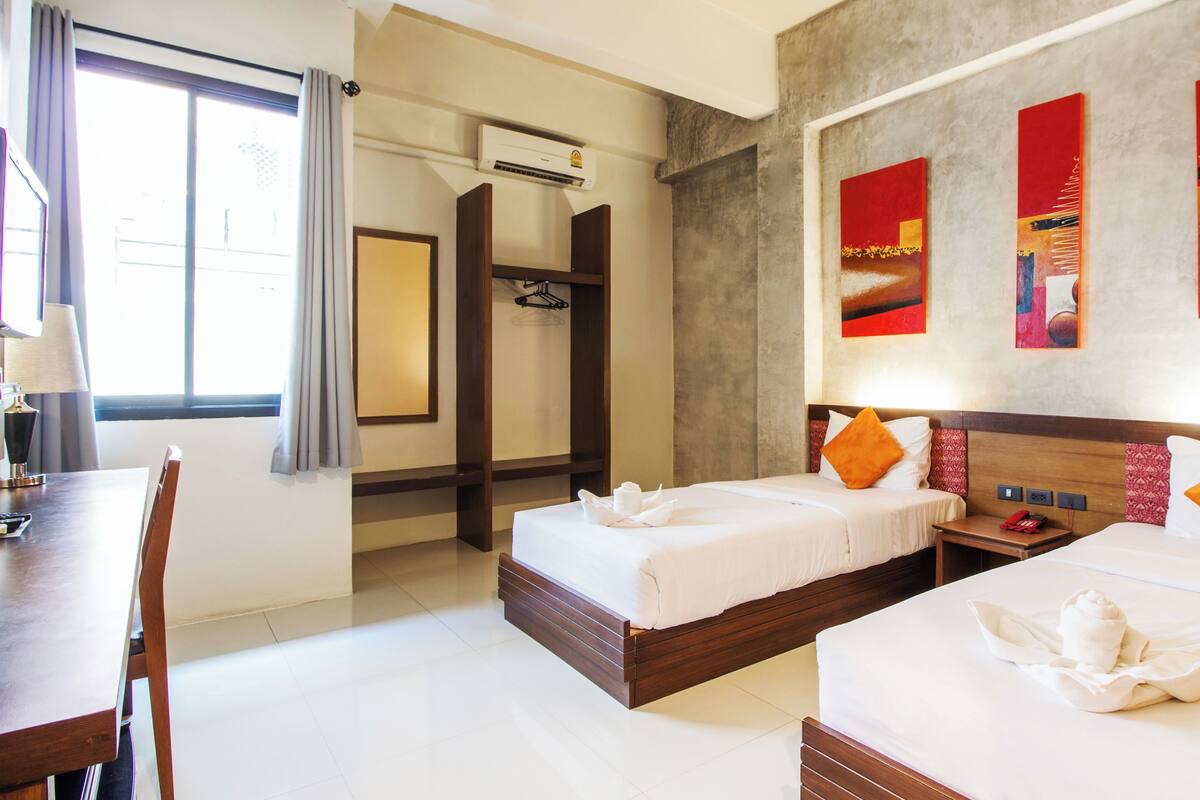 A hotel room with two beds, a desk, and a chair. There is a window with curtains, an air conditioning unit, and artwork on the wall.