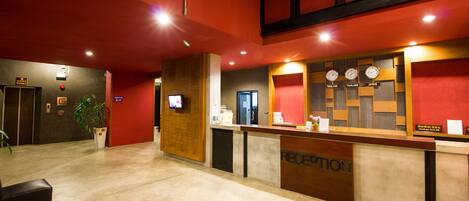 A hotel reception area with a reception desk, a TV screen, a clock displaying time in three different countries, and a sign indicating the check-out time.