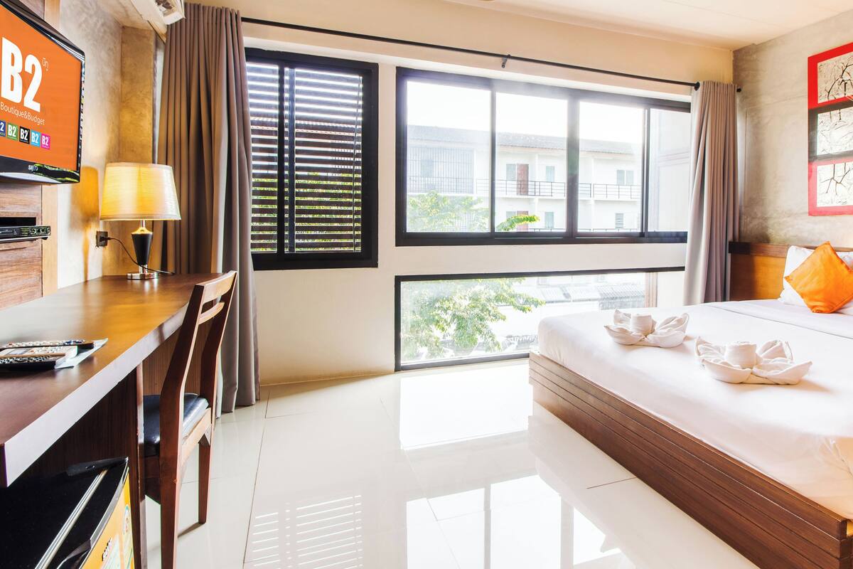 A hotel room with a large bed, a desk with a lamp, a TV, and a balcony with a view of buildings.