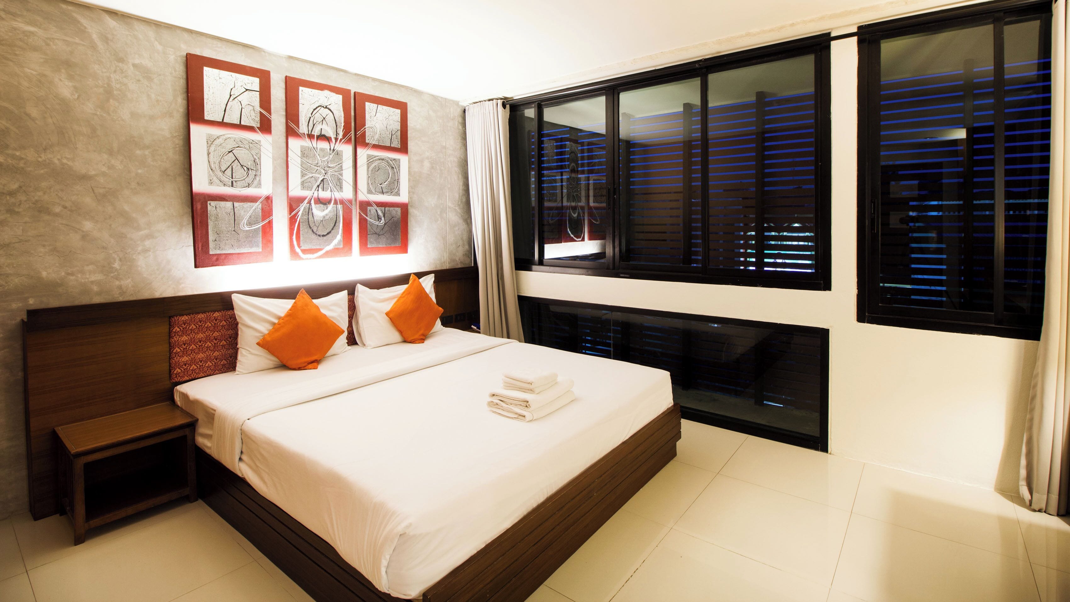 Una habitación de hotel con una cama grande, una mesita de noche, una ventana con persianas y una pared decorada con cuadros.