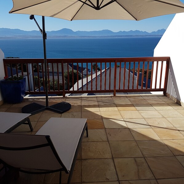 A balcony with a view of the sea and mountains, a table, chairs, and a large umbrella.