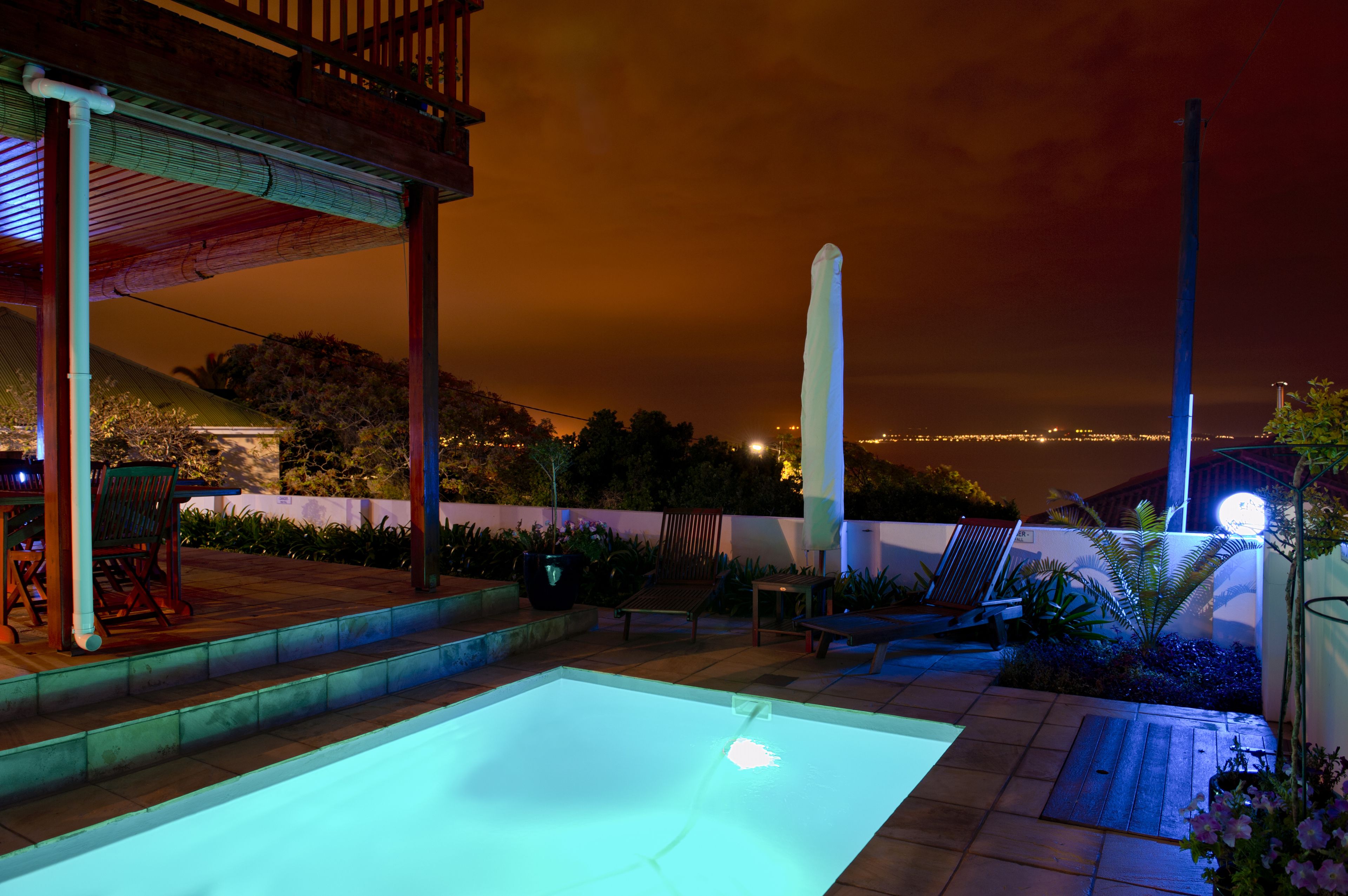 A swimming pool with a wooden deck and lounge chairs, surrounded by a landscaped garden and a view of a cityscape at night.